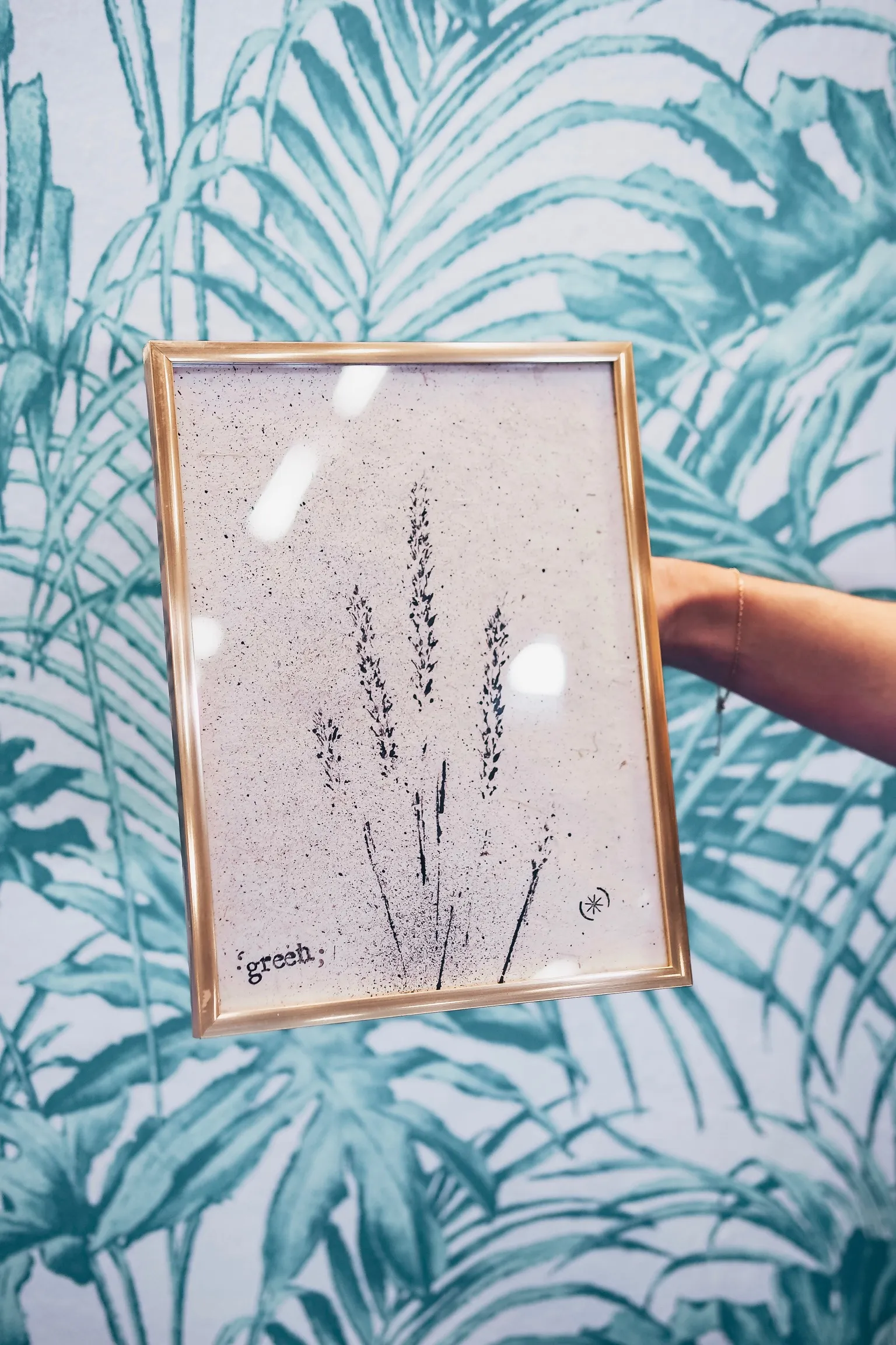 Tableau végétal DIY avec empreintes de lavande dans un cadre doré tenu devant un papier peint feuillage