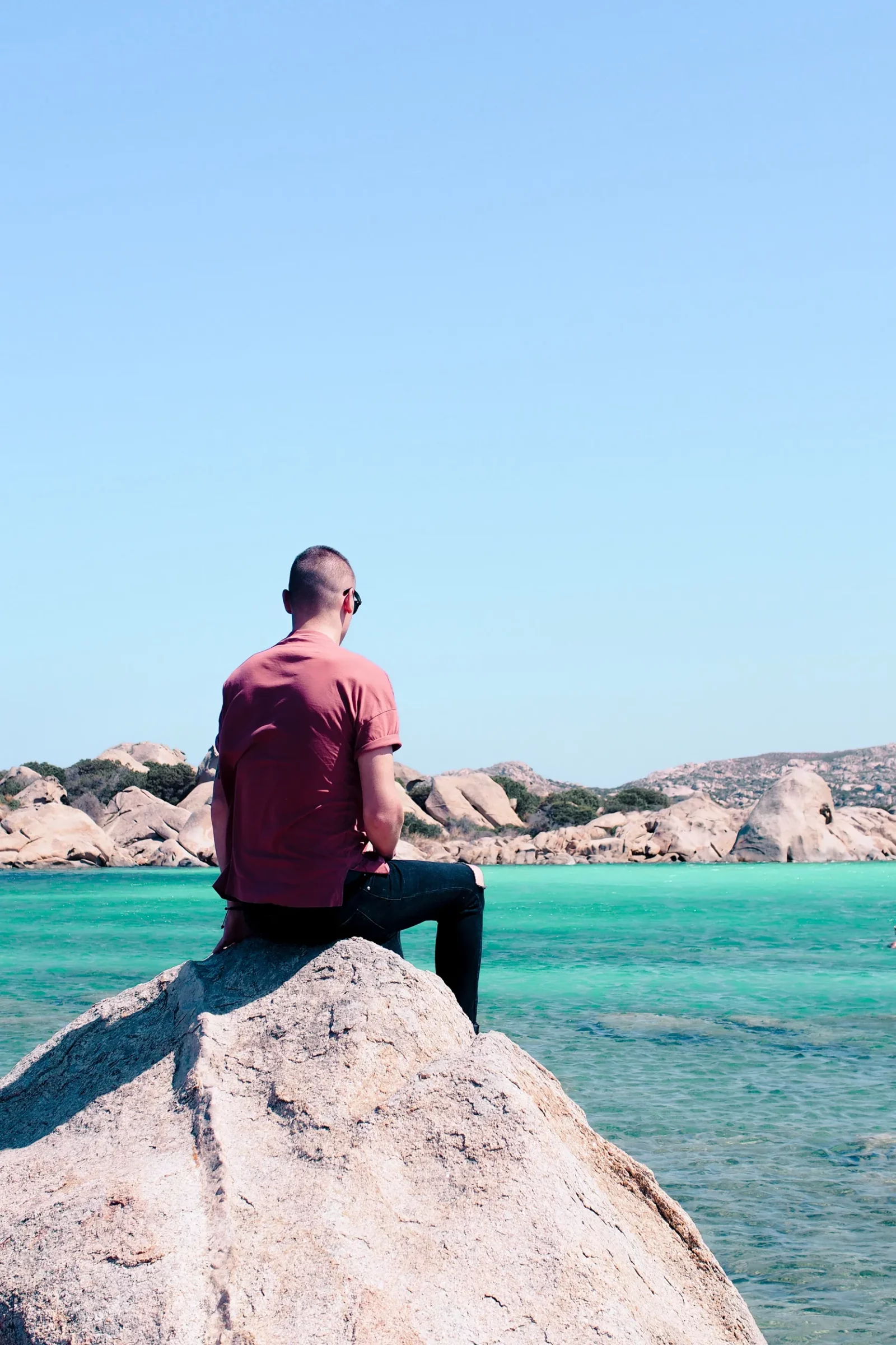 Voyageur assis sur un rocher de granit face à l'eau turquoise de l'archipel de La Maddalena